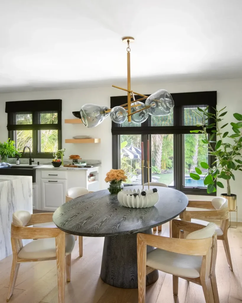 Modern dining area with a round black wood table, light oak chairs, sculptural brass chandelier with glass globes, and black natural shades over large windows overlooking a lush backyard pool.