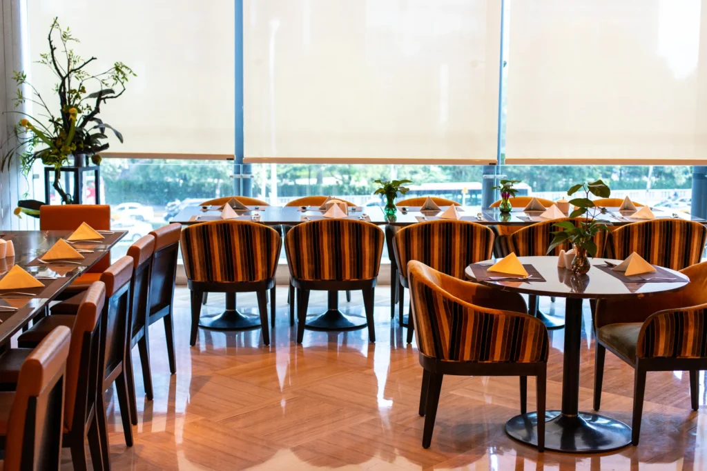 Modern restaurant dining area with neatly set tables and upholstered chairs, featuring large windows fitted with motorized roller shades that filter natural light.