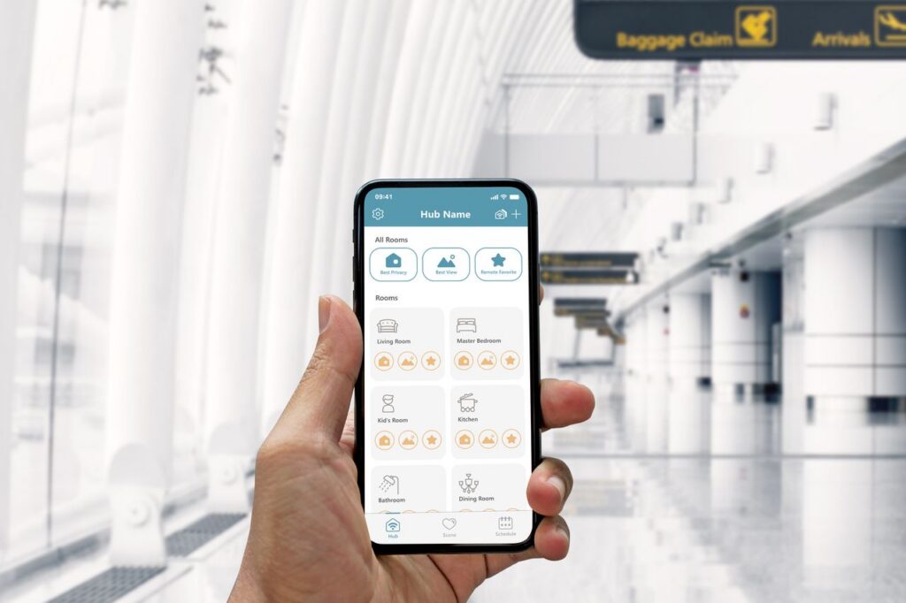 Hand holding a smartphone displaying a smart home app controlling motorized roller shades across multiple rooms, set against a bright, modern airport terminal background.