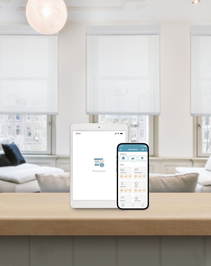 Smartphone and tablet displaying a smart home app used to control motorized roller shades, set on a kitchen counter in a bright, modern living space featuring installed motorized roller shades across multiple windows.