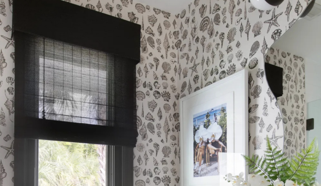 Black woven Roman shade over small window in powder bathroom with black-and-white shell wallpaper, white sink, black faucet, and modern decor.