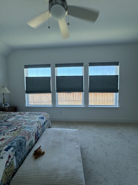 Top down bottom up Roman shades in a Buda, TX bedroom with three large windows, allowing natural light through the top while maintaining privacy below with custom gray fabric shades.