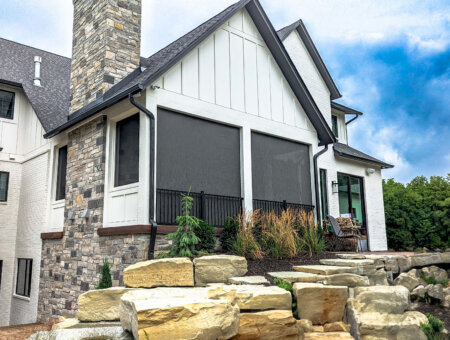 Exterior shades on a covered patio in Austin, TX with white siding, stone accents, black railing, and landscaped rock steps.